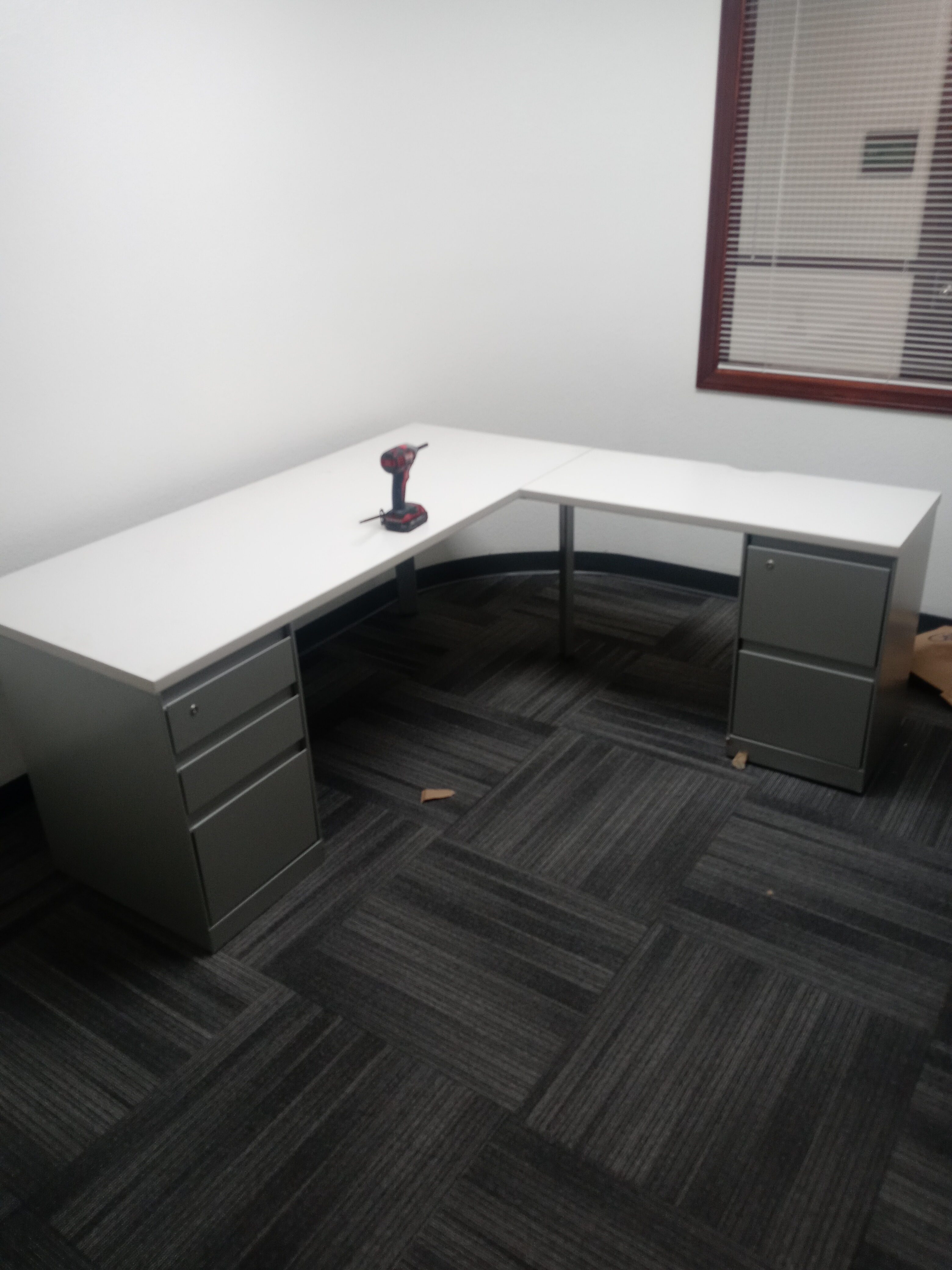 L-shaped office desk with a white tabletop and gray metal drawer units, set up in a professional workspace with a cordless drill placed on top