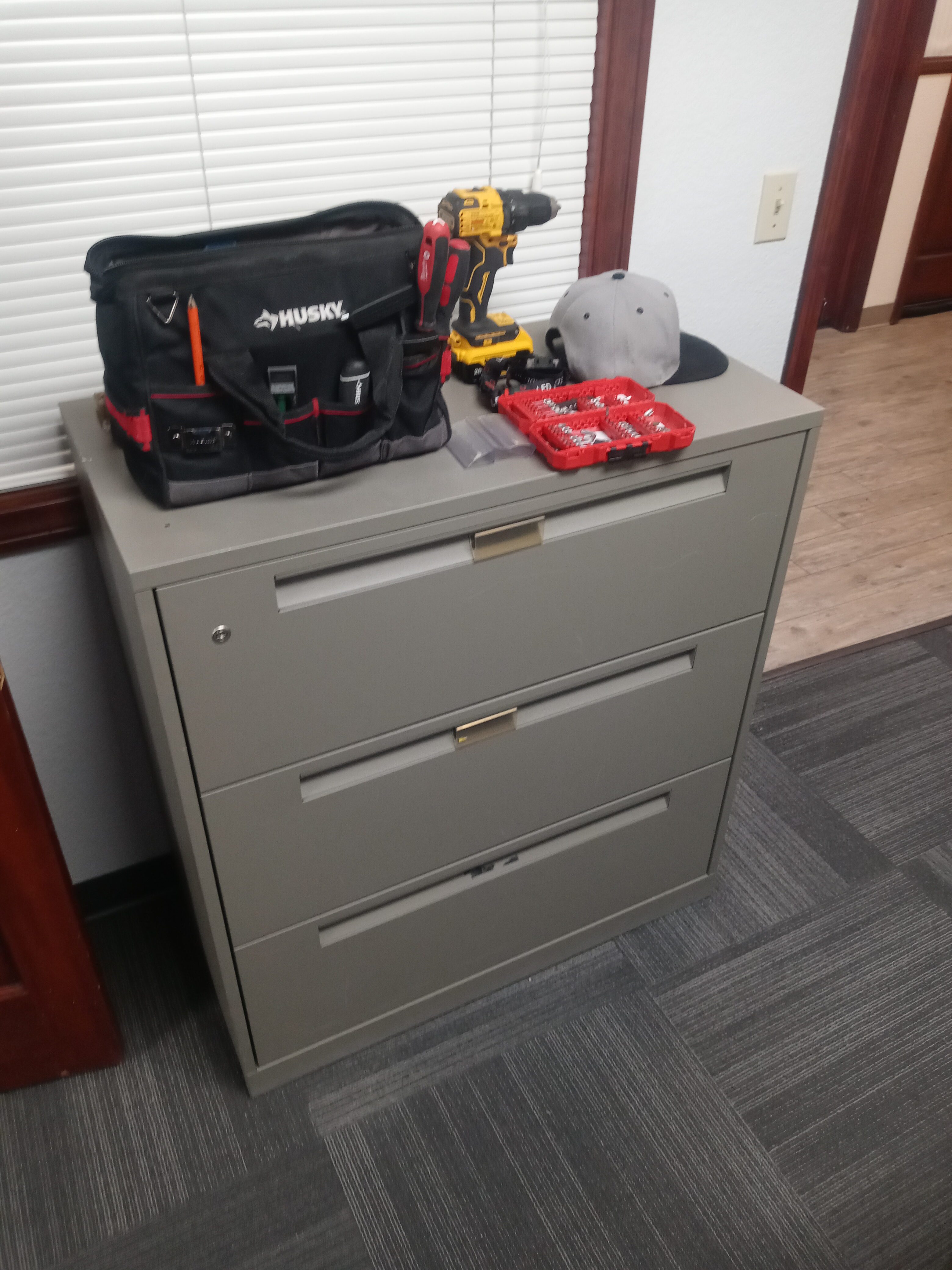 Gray metal filing cabinet with a Husky tool bag, a cordless drill, a baseball cap, and an organized drill bit set placed on top in an office setting.