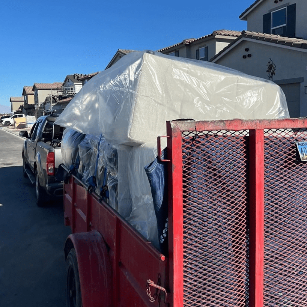 A red trailer carrying a securely wrapped mattress and furniture, safely transported in a residential neighborhood