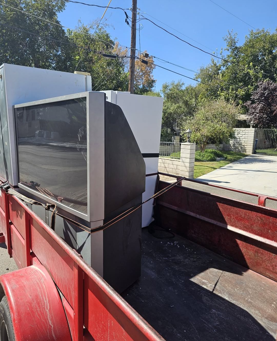 Red trailer loaded with discarded appliances and an old projection TV, secured with straps, ready for junk removal in a residential neighborhood