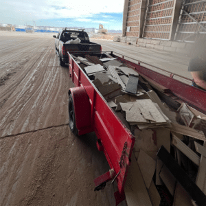 Red trailer loaded with construction debris, including wooden planks and broken tiles, hitched to a pickup truck at an industrial site.