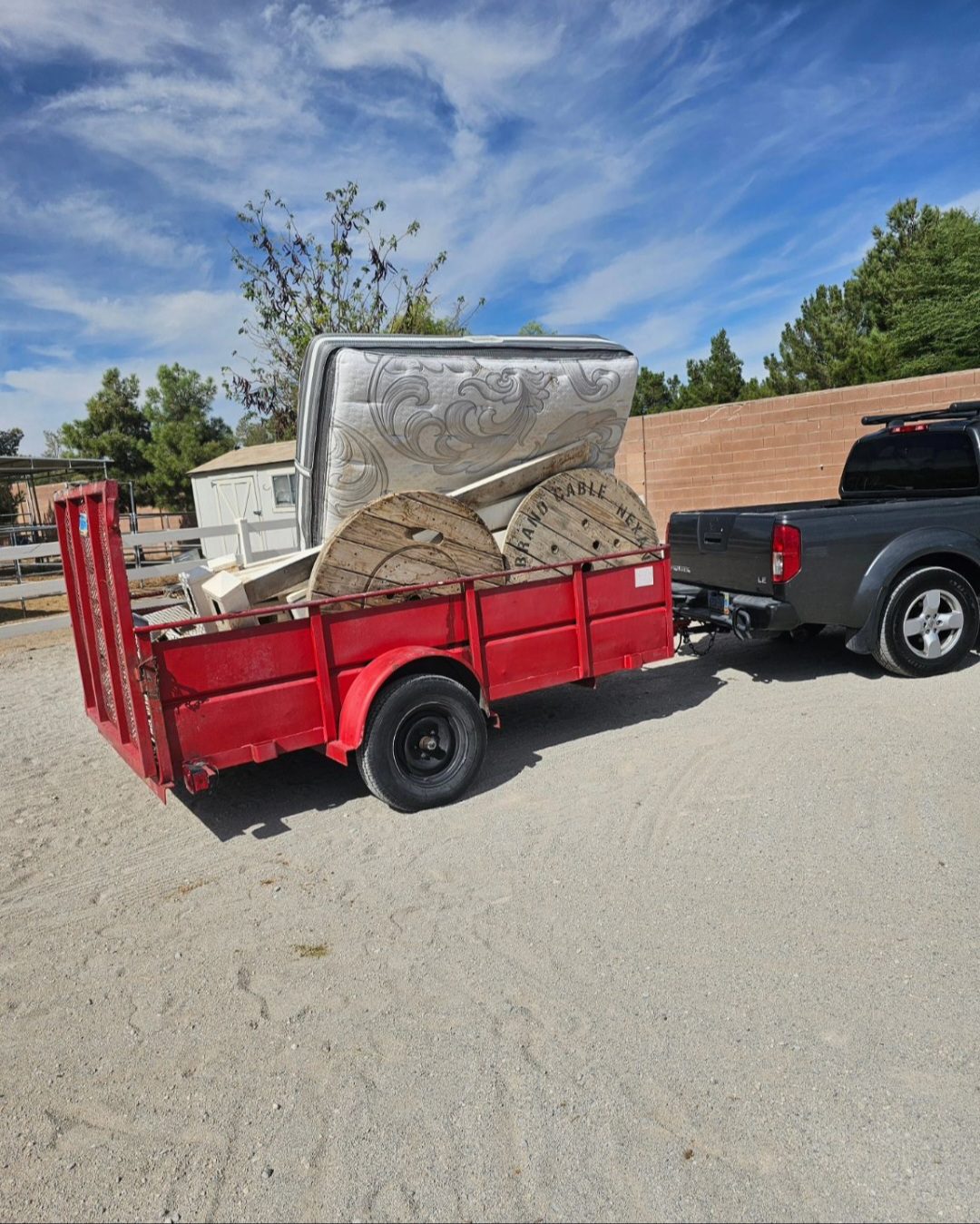 Red trailer attached to a gray pickup truck loaded with discarded items, including a mattress, wooden cable spools, and miscellaneous debris in an outdoor setting.
