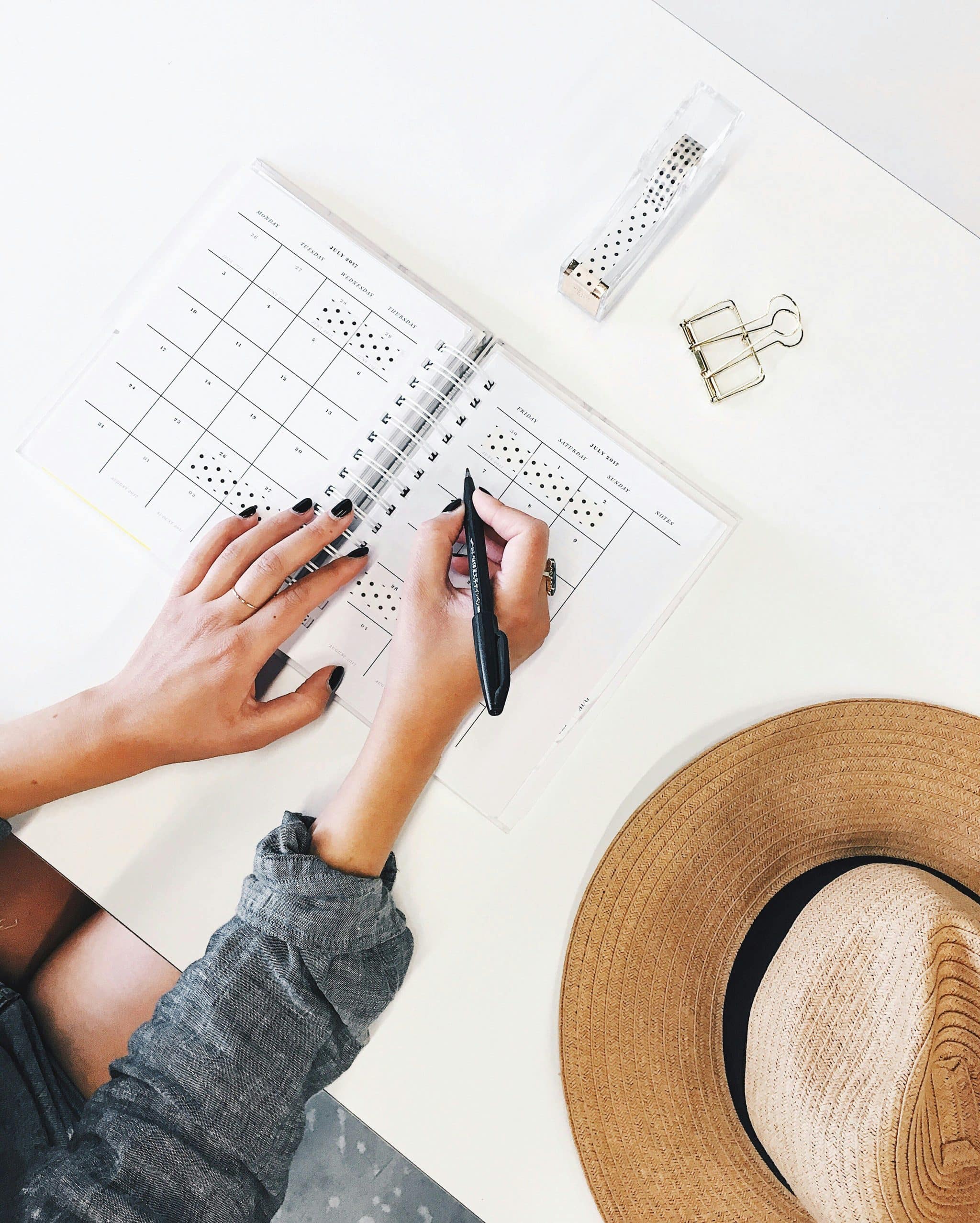 Close-up of hands writing in a planner, organizing schedules, with a straw hat and office supplies on a white desk