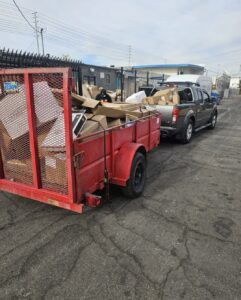 A loaded red trailer attached to a pickup truck, filled with discarded cardboard boxes and other junk, ready for removal in an industrial area